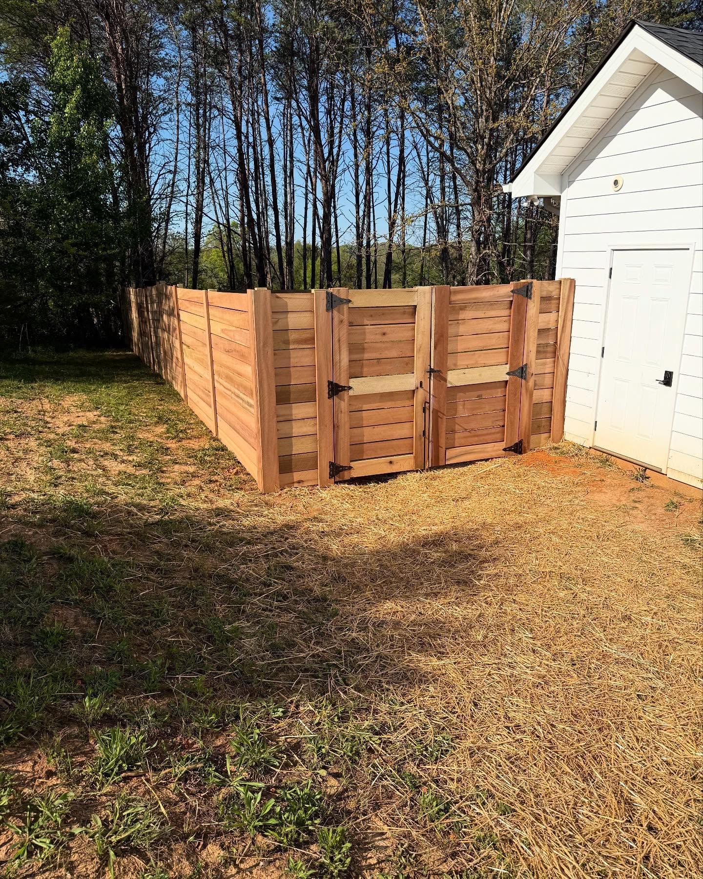 Wooden compost bin enclosure beside a white shed in a wooded yard