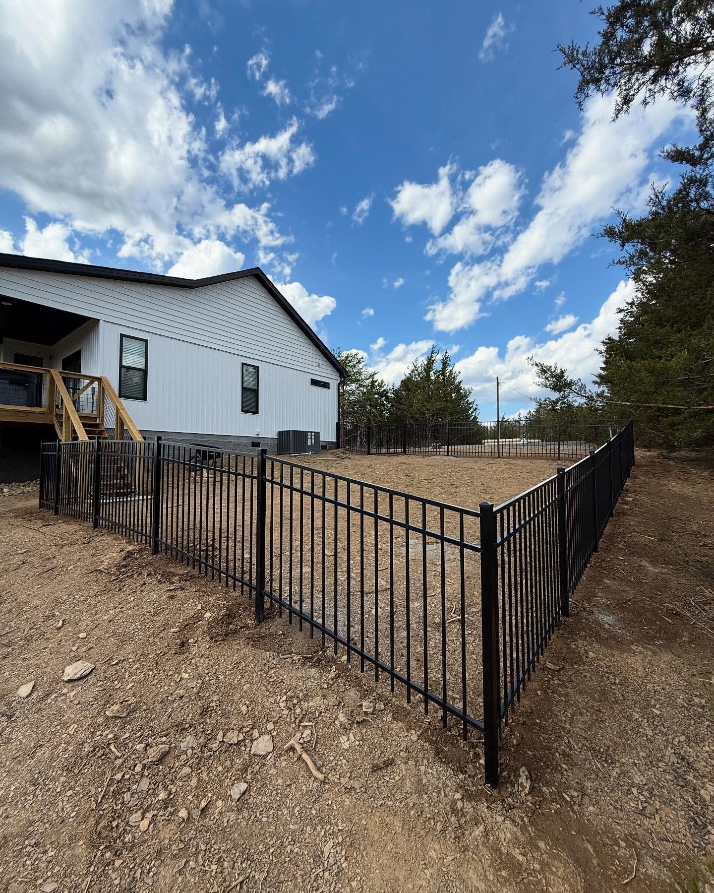 Black metal fence enclosing gravel yard beside a white house under a blue, cloudy sky