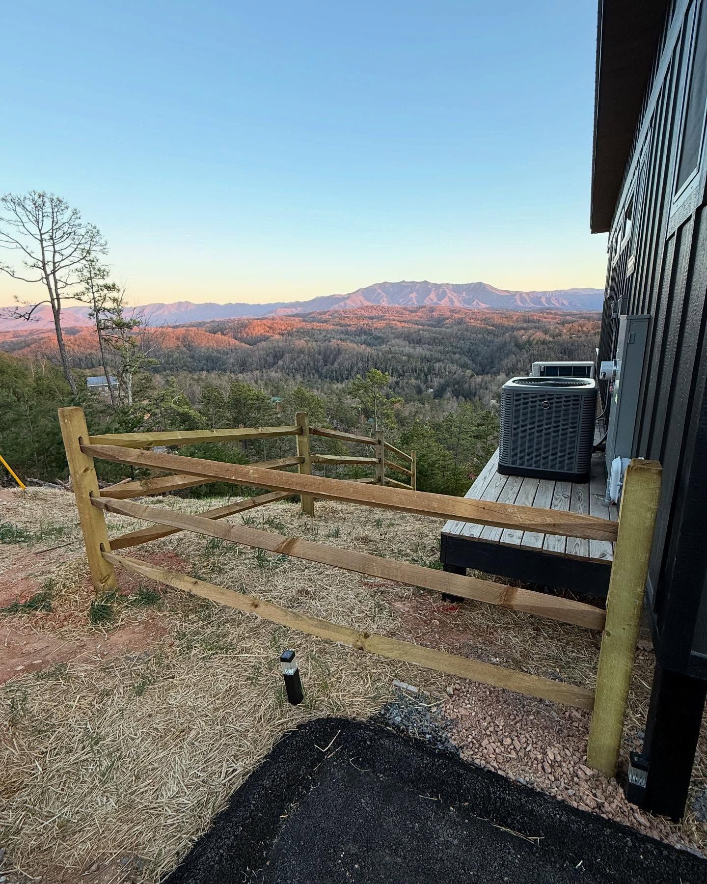 Wooden deck overlooking a sunset-lit mountain valley beside a house, with an AC unit and bare trees.