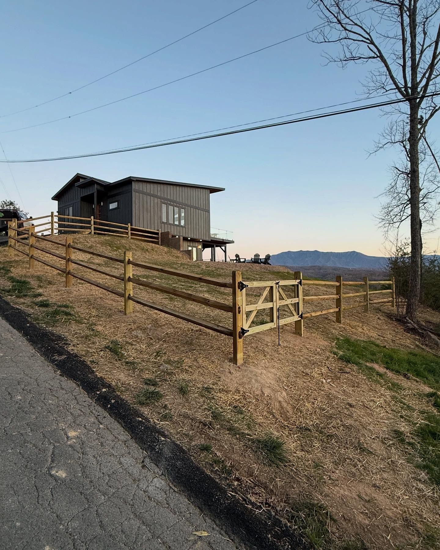 Rural hillside with a small dark cabin, wooden fence, gate, and distant mountains under a clear sky