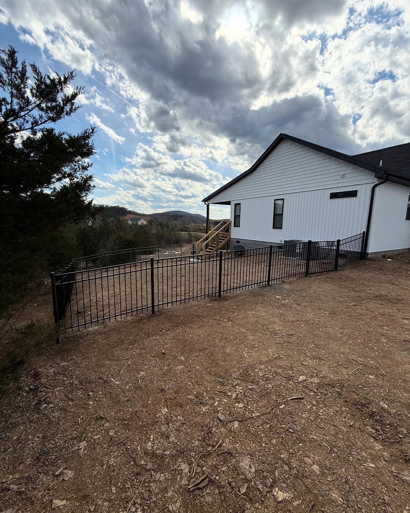 White house with black roof beside a fenced gravel yard under a cloudy sky, with hills in the distance