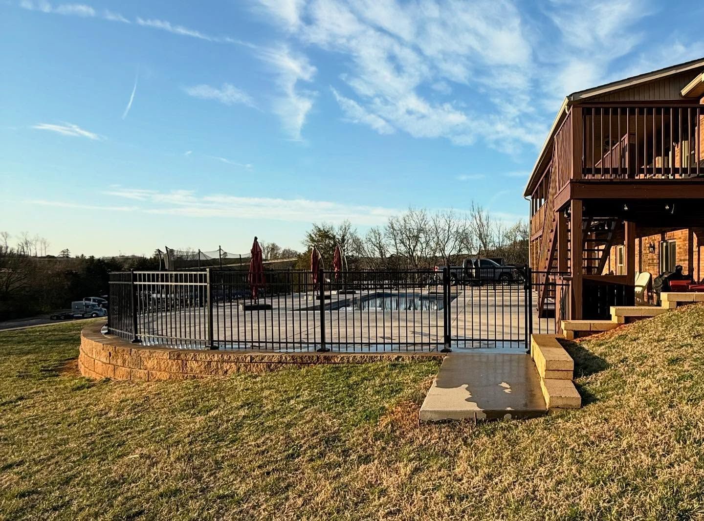 Backyard pool with black safety fence beside a wooden house at sunset