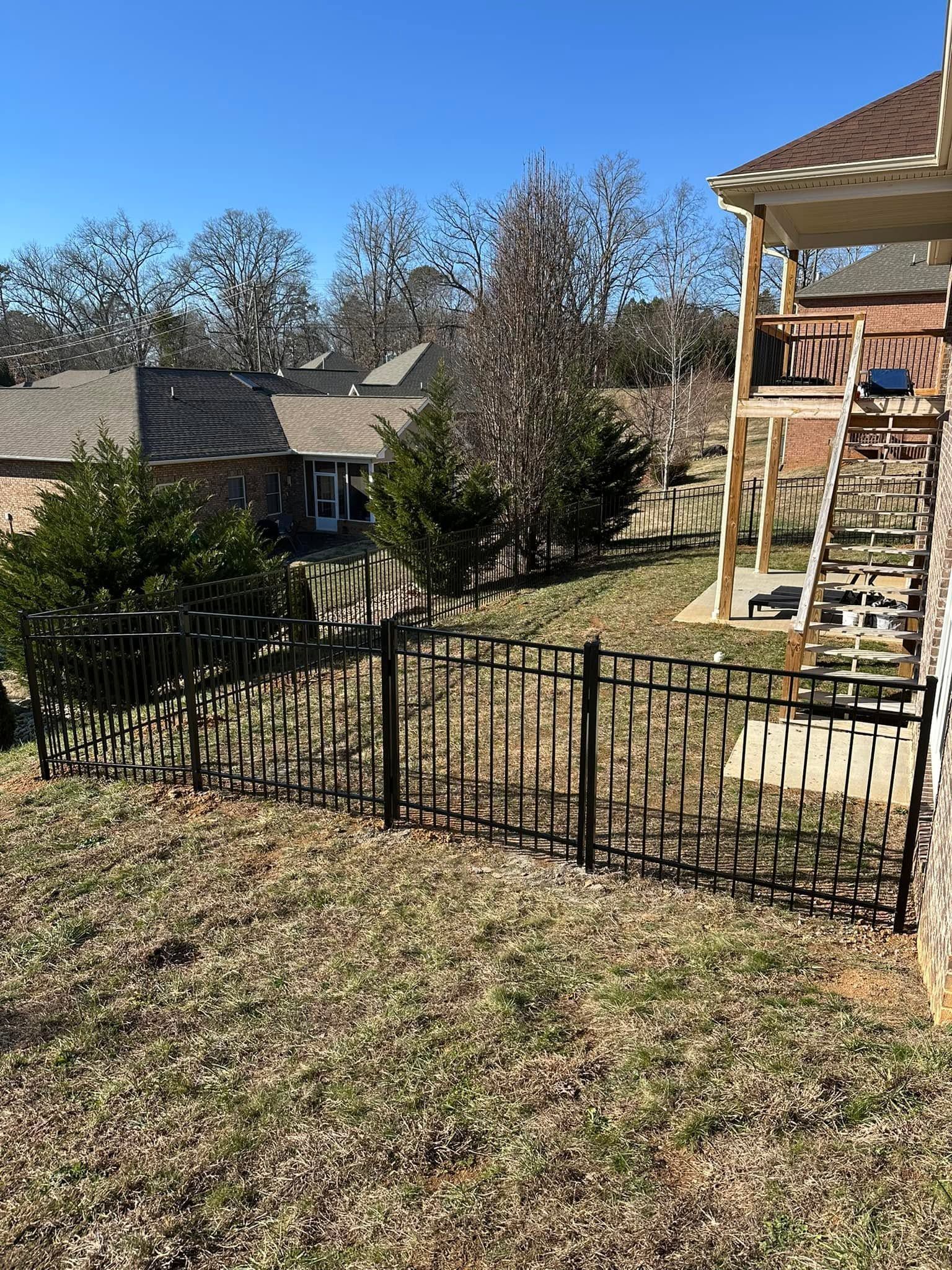 Backyard with black metal fence, trees, and a stone house patio with stairs and chairs under a clear blue sky