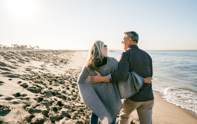 Couple walking arm-in-arm along a sandy beach at sunset, with waves on the shore and dunes nearby
