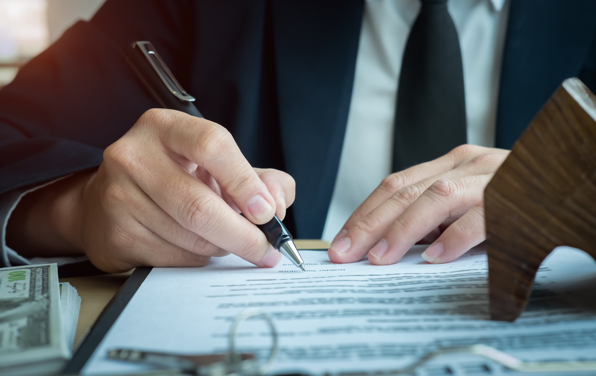Hands signing a document beside a pen and cash on a desk