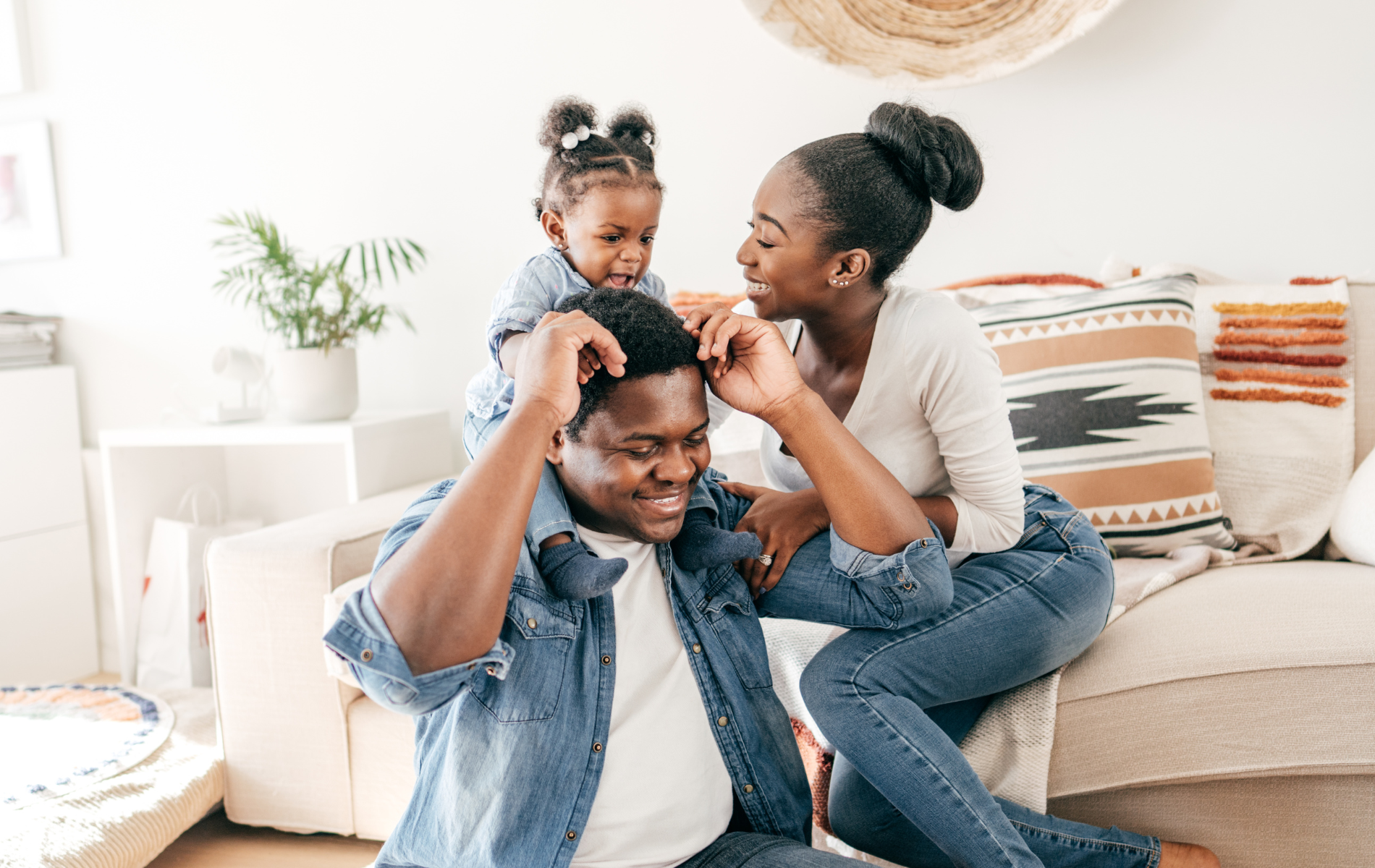 Family laughing together on a couch, with a child playfully perched on an adult's shoulders.