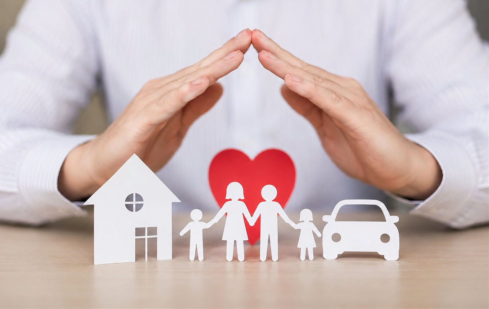 Hands forming a protective roof over a family, house, heart, and car on a table