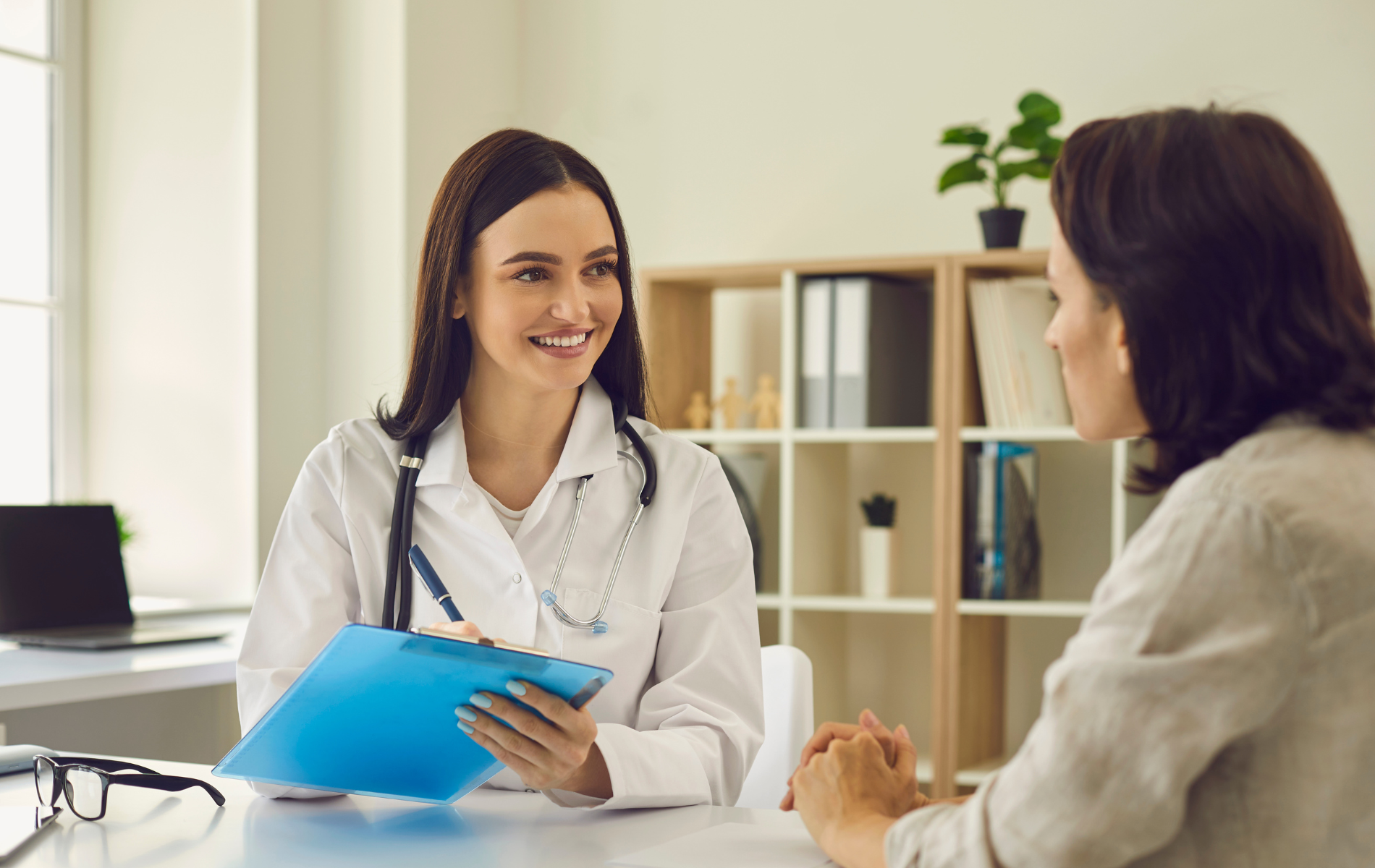 Doctor speaking with a patient at a desk, holding a blue clipboard in a bright office.