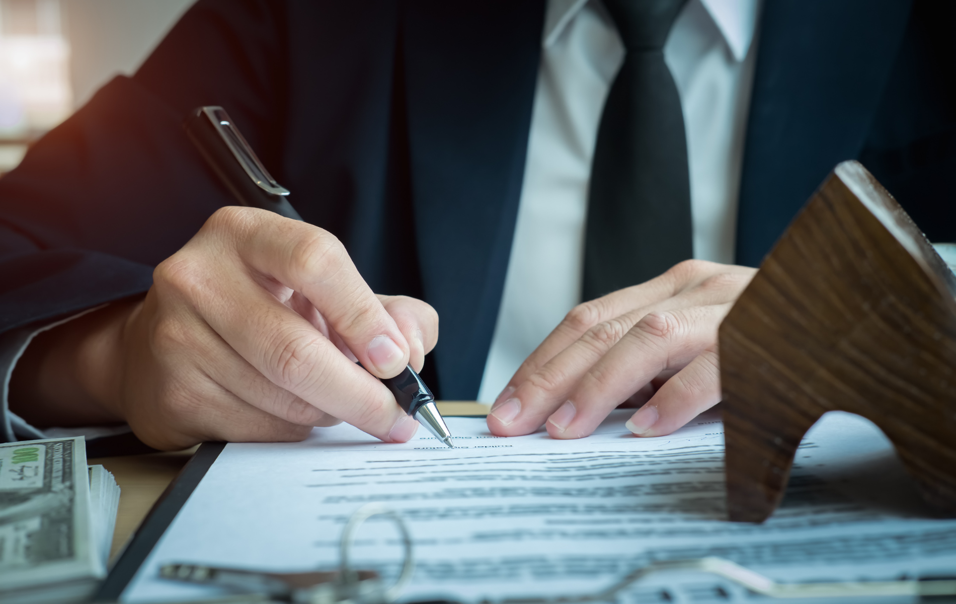 Person signing a document with a pen beside a wooden gavel and stacks of money