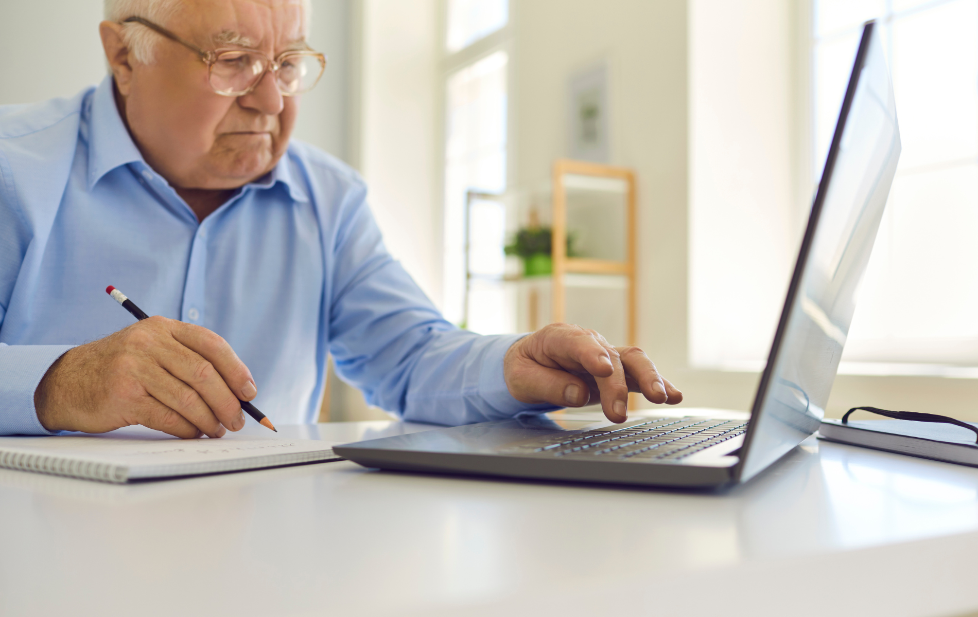 Older man writing in a notebook and using a laptop at a bright desk