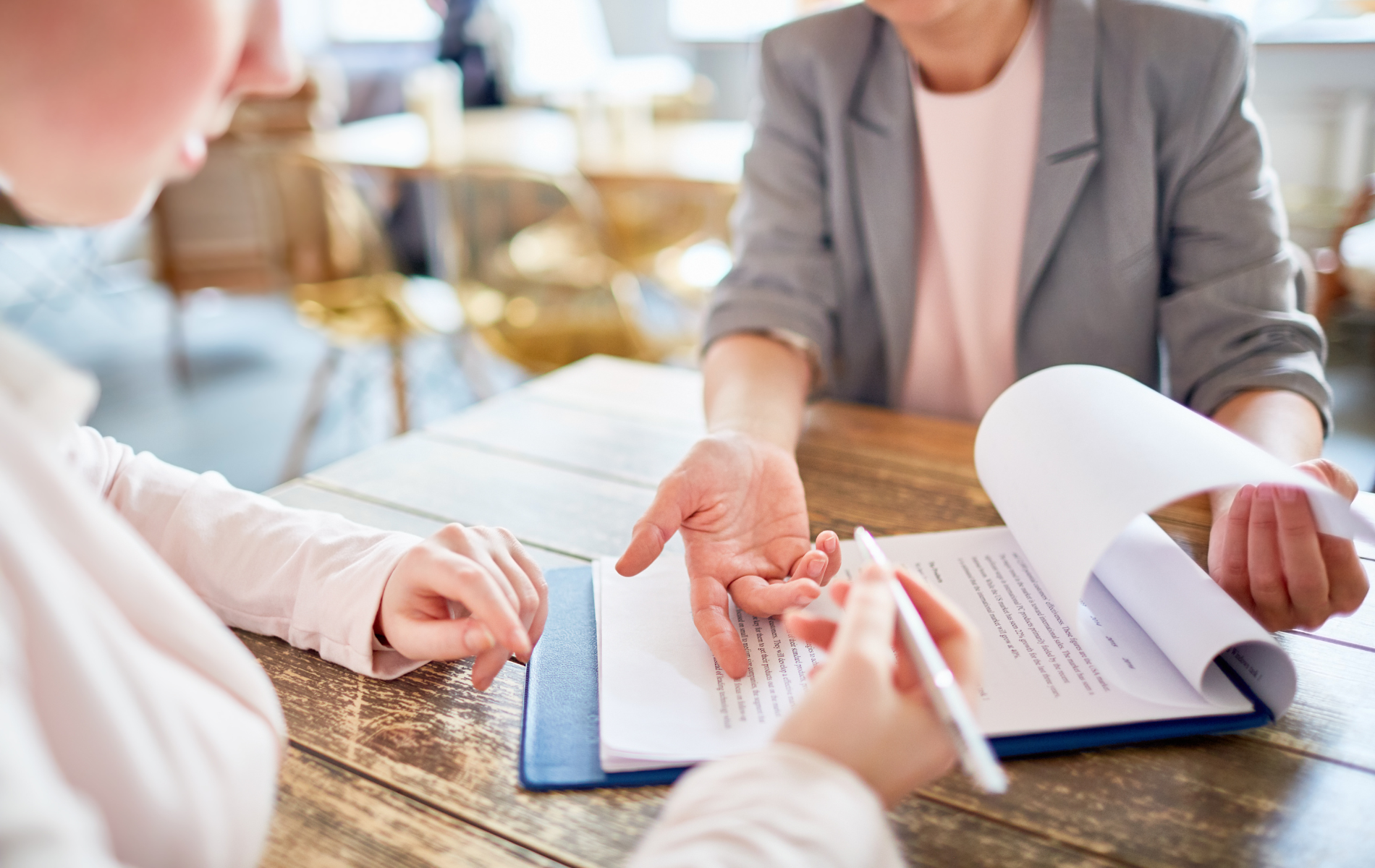 People in a meeting exchanging documents over a wooden table, with a notebook and pen visible.