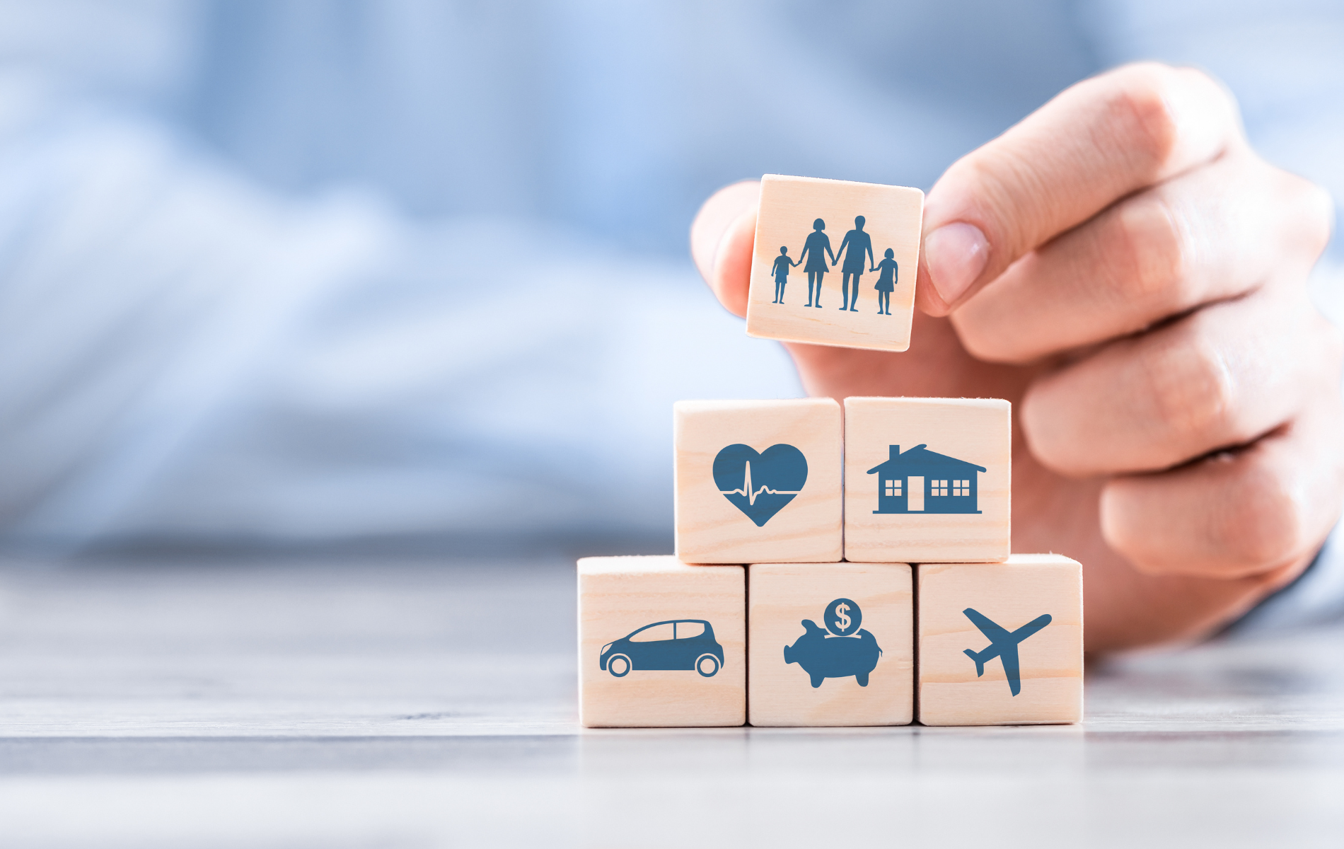 Hand arranging stacked wooden blocks with icons for family, health, home, car, piggy bank, and travel