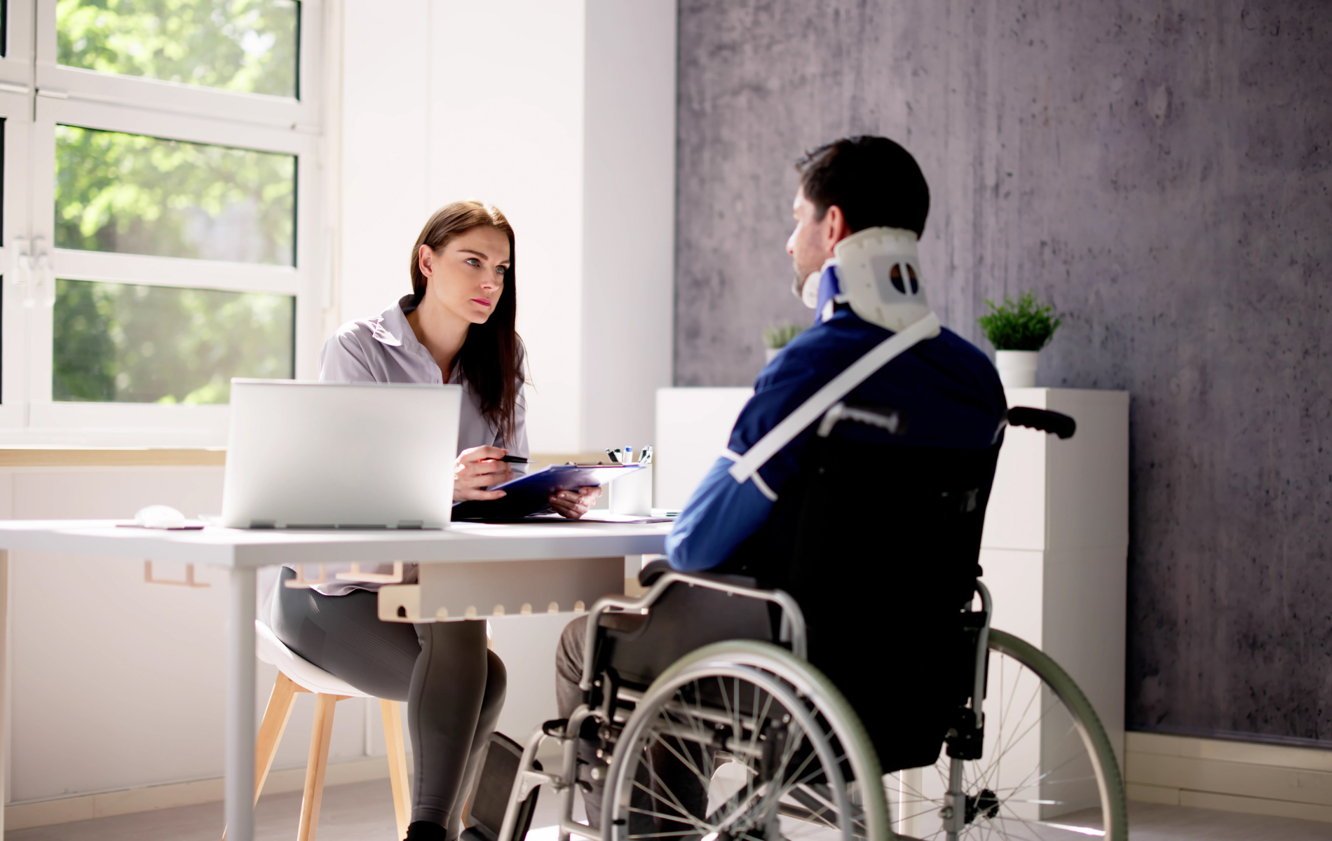 Two coworkers meeting at a table, one in a wheelchair, in a bright office with a laptop.