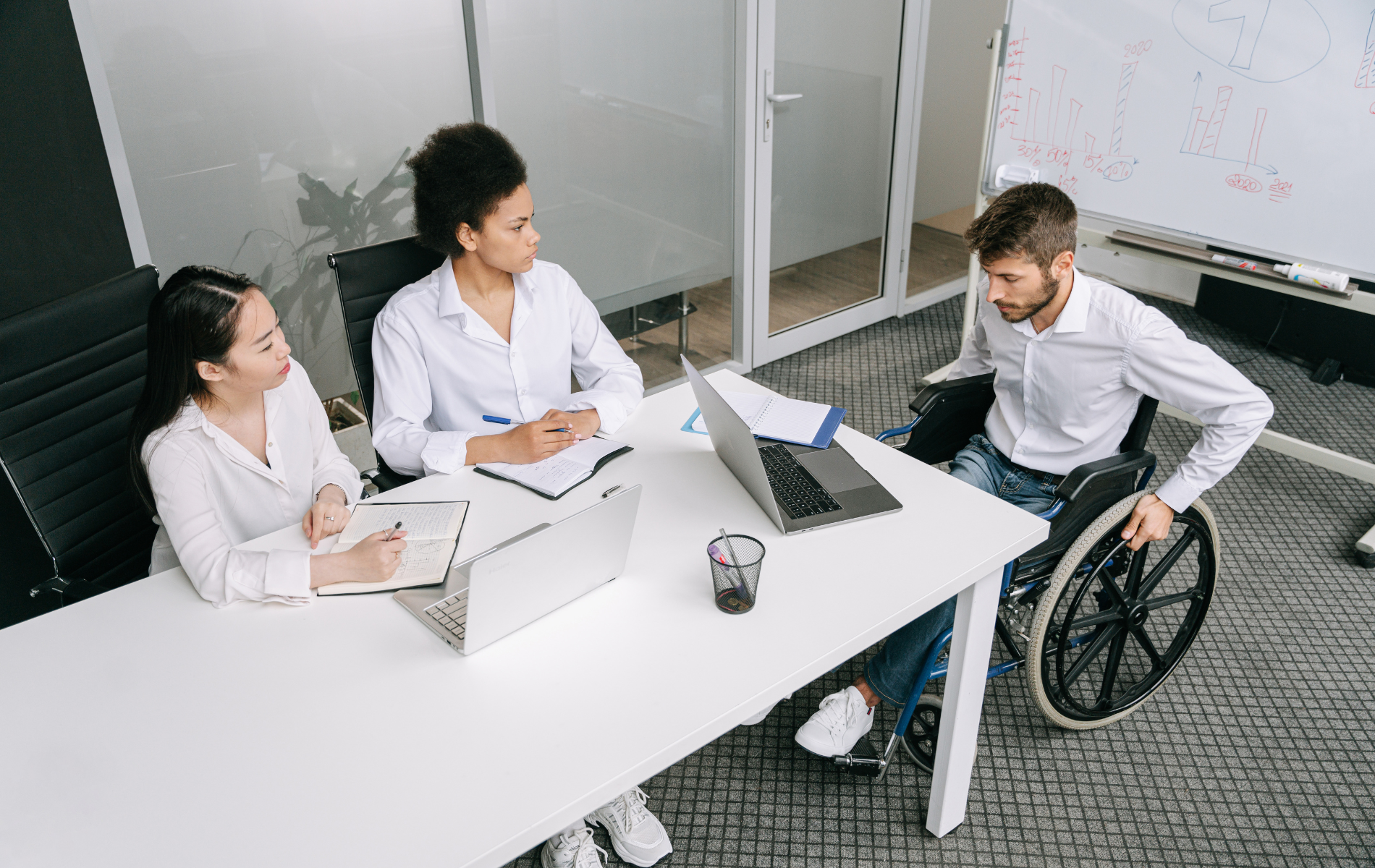 Three coworkers in a meeting room, one in a wheelchair, discussing documents around a white table.