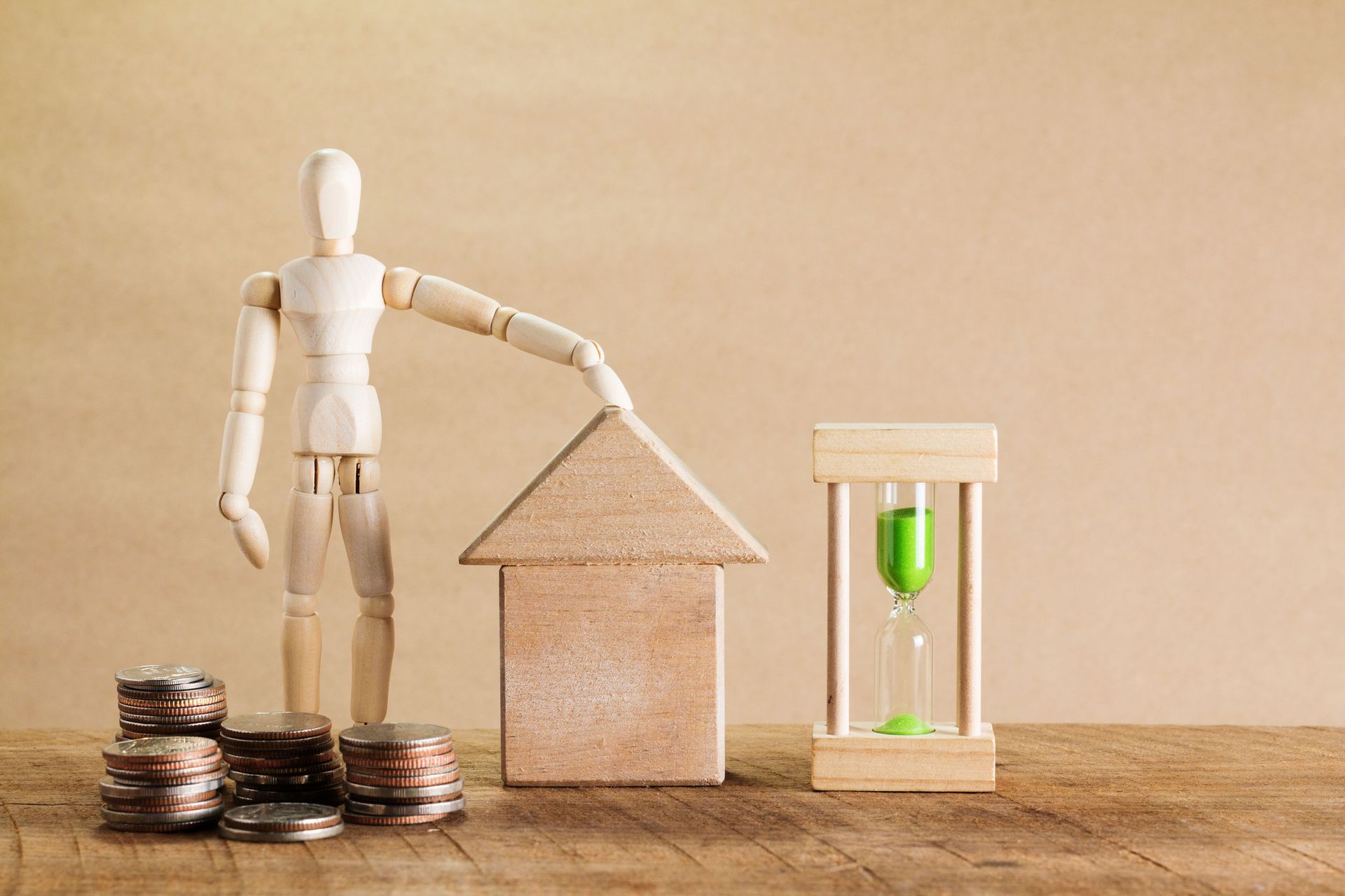 Wooden figure with house model, coin stacks, and hourglass with green sand on a table