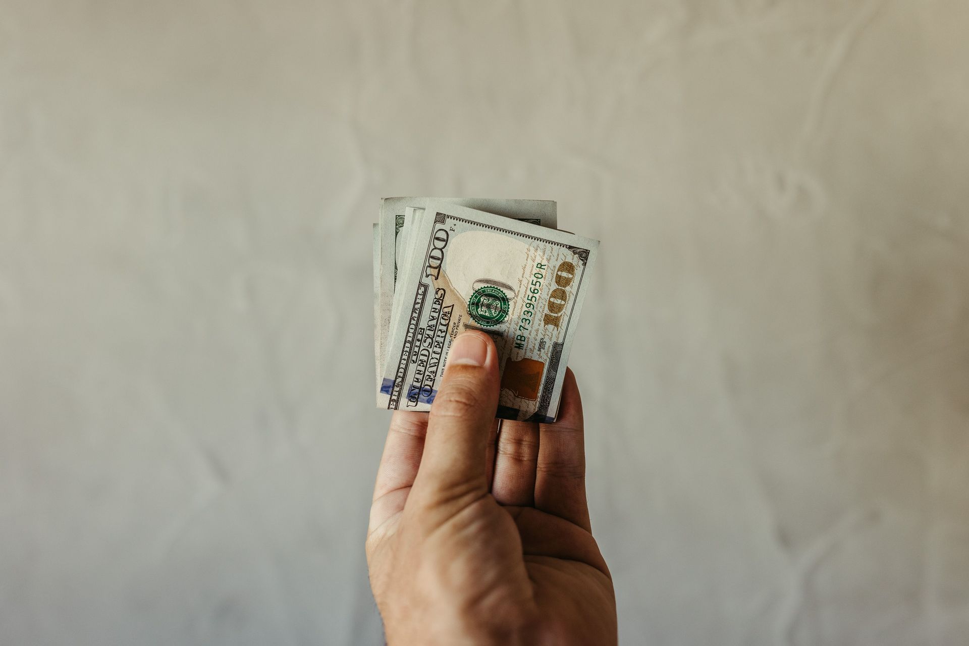 Hand holding a small stack of U.S. dollar bills against a plain wall.