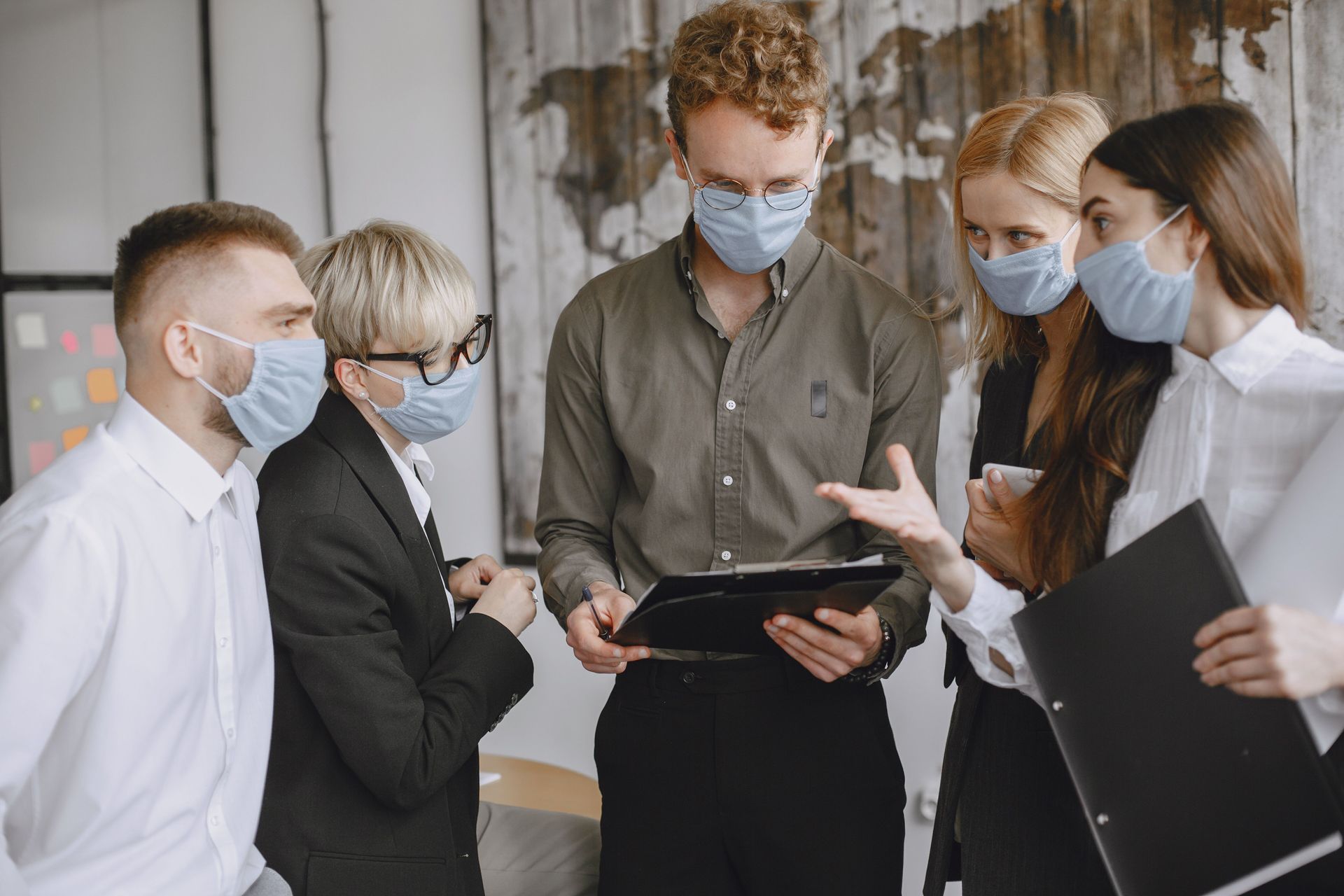 Masked coworkers in a meeting, reviewing a tablet and documents in an office.