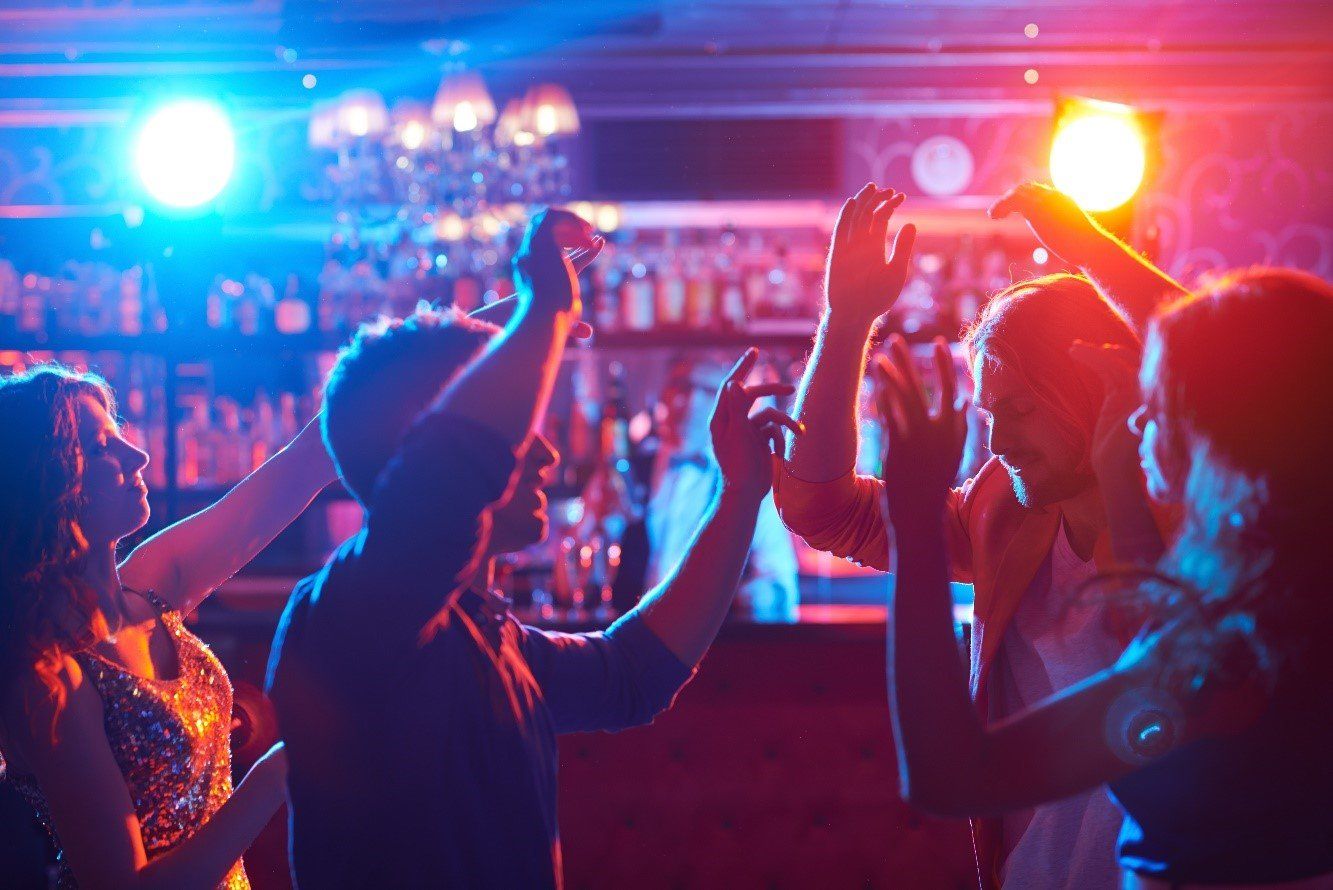 A group of people dancing in a dimly lit bar, silhouetted against vibrant blue and red stage lights.
