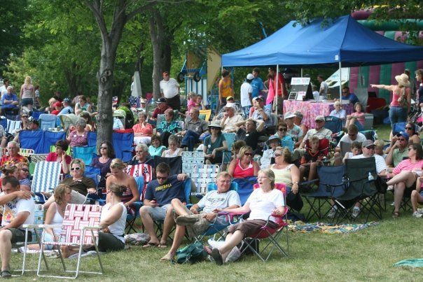 A crowd of people seated in folding chairs on a grassy lawn near trees, with a blue tent in the background.