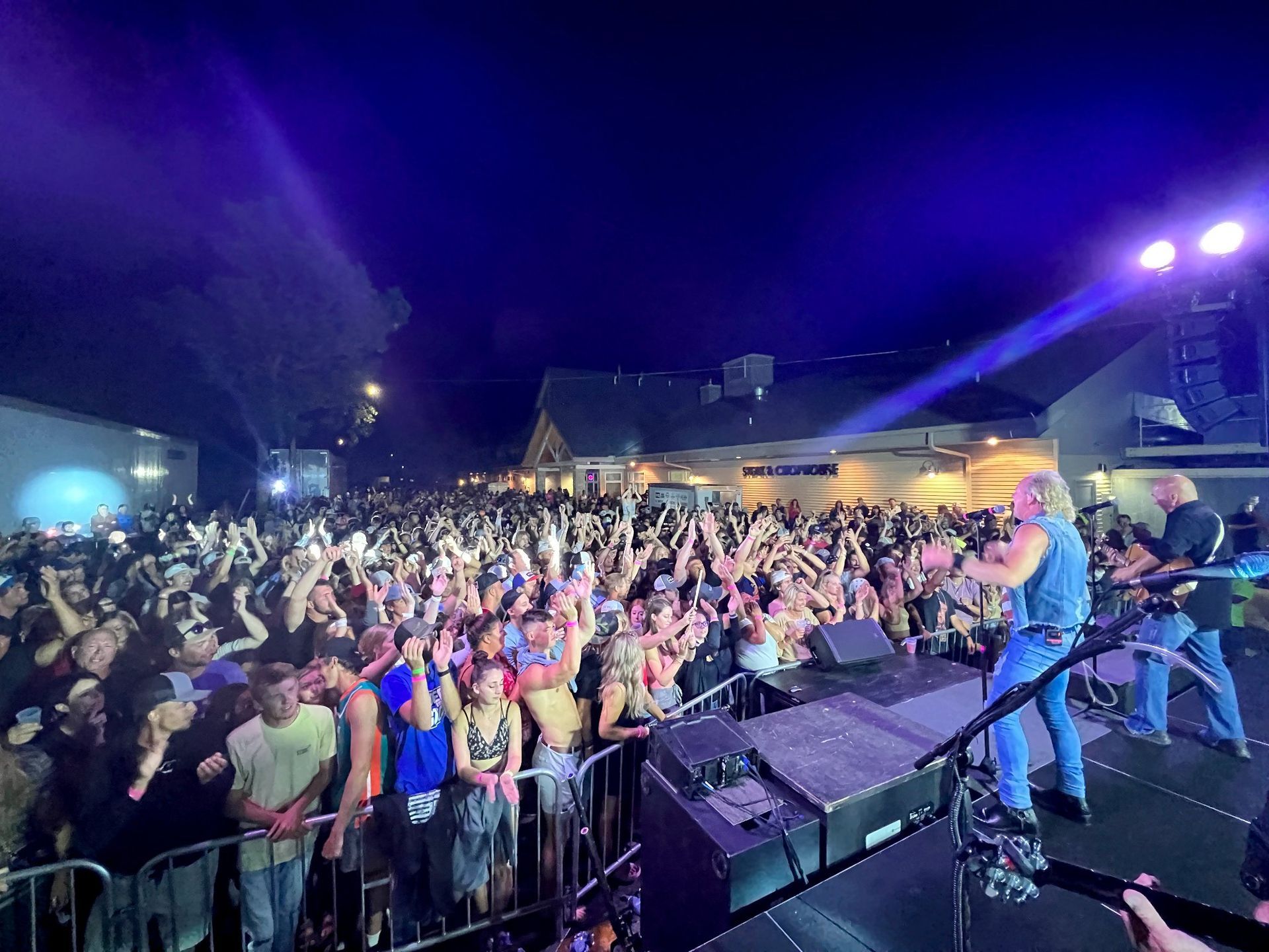 A band performs on an outdoor stage at night before a large, cheering crowd with hands raised.