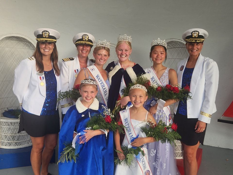 A group of pageant winners and official escorts wearing nautical-themed uniforms and crowns stand together indoors.