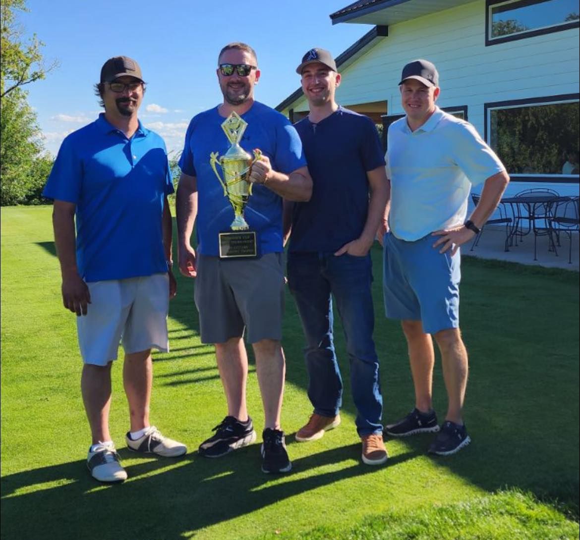 Four people stand in a sunny, grassy yard posing with a gold trophy, wearing casual clothes and baseball caps.