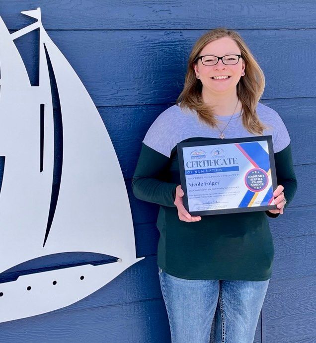 A smiling person wearing glasses and a two-toned shirt holds an award certificate against a blue wall with a boat cutout.