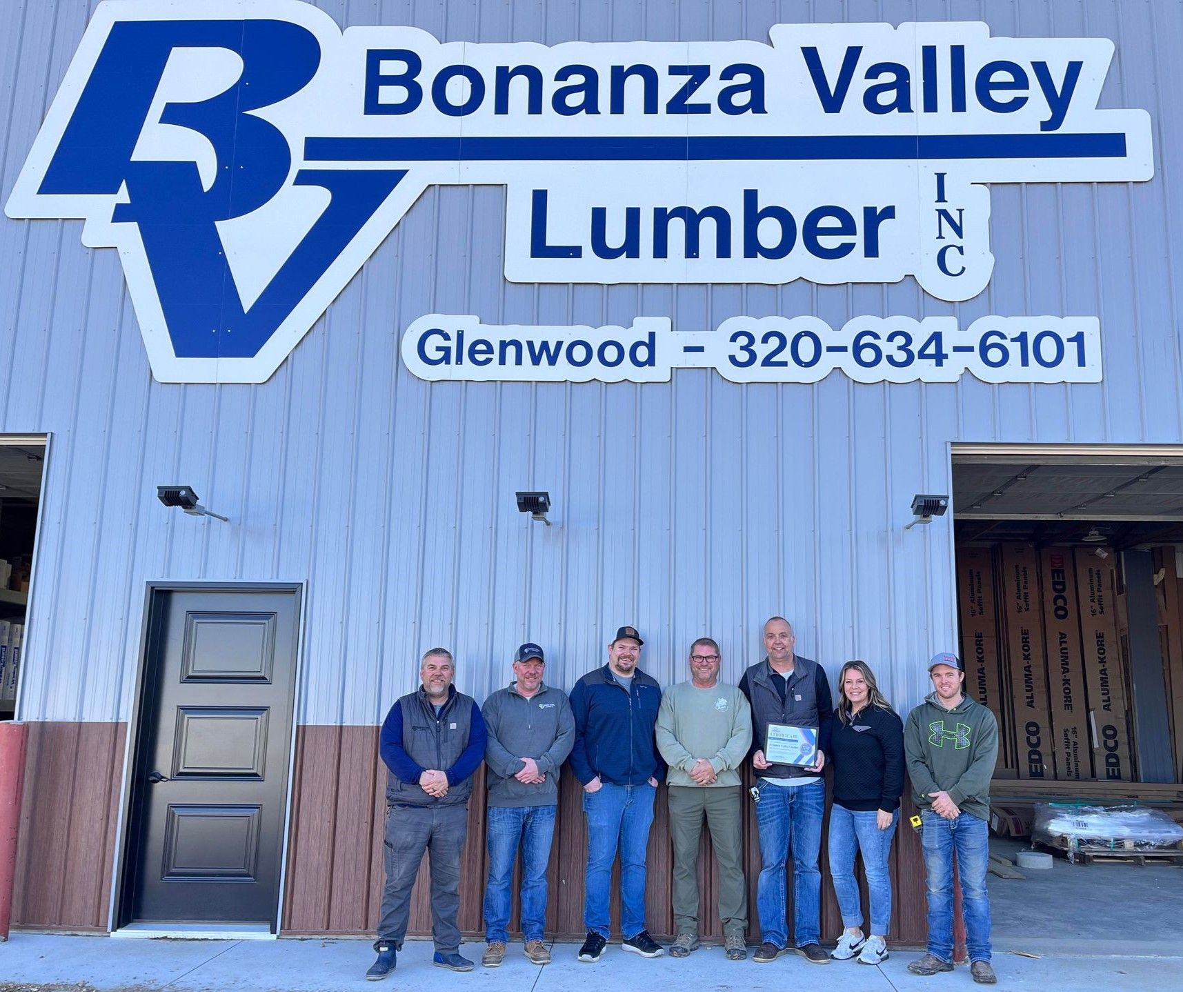 A group of seven people stands in front of a metal building marked with the Bonanza Valley Lumber sign in Glenwood.