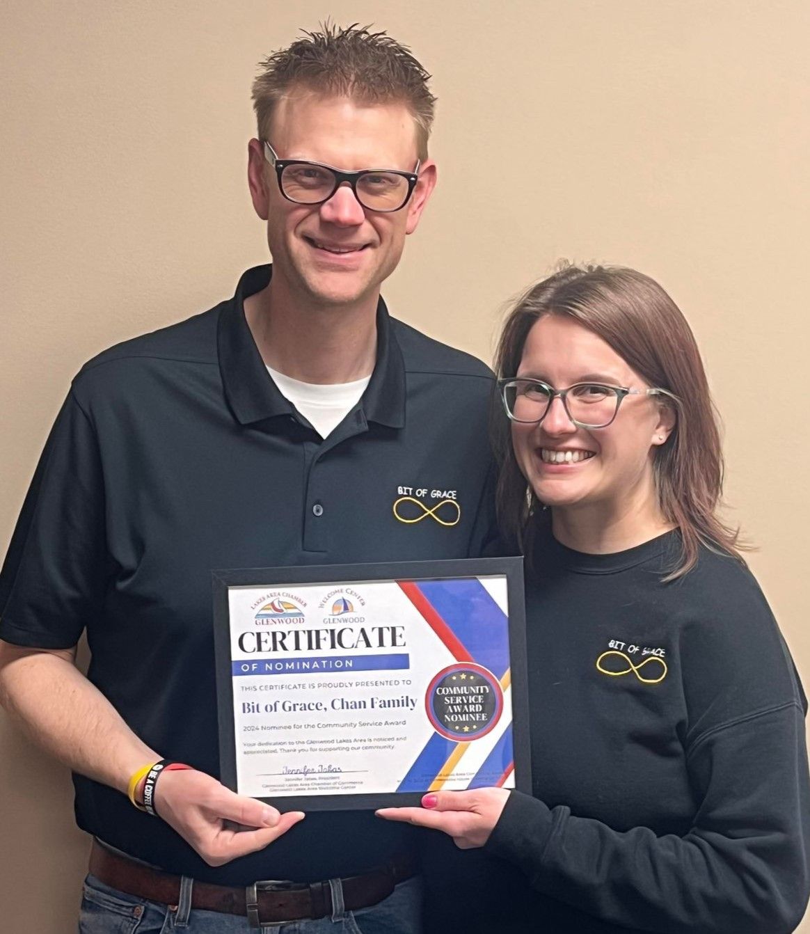 Two people smiling while holding a framed certificate against a plain wall.