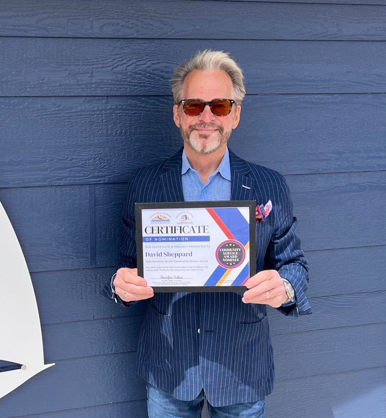 A person wearing sunglasses and a patterned navy blazer holds a framed certificate in front of a blue horizontal-slat wall.
