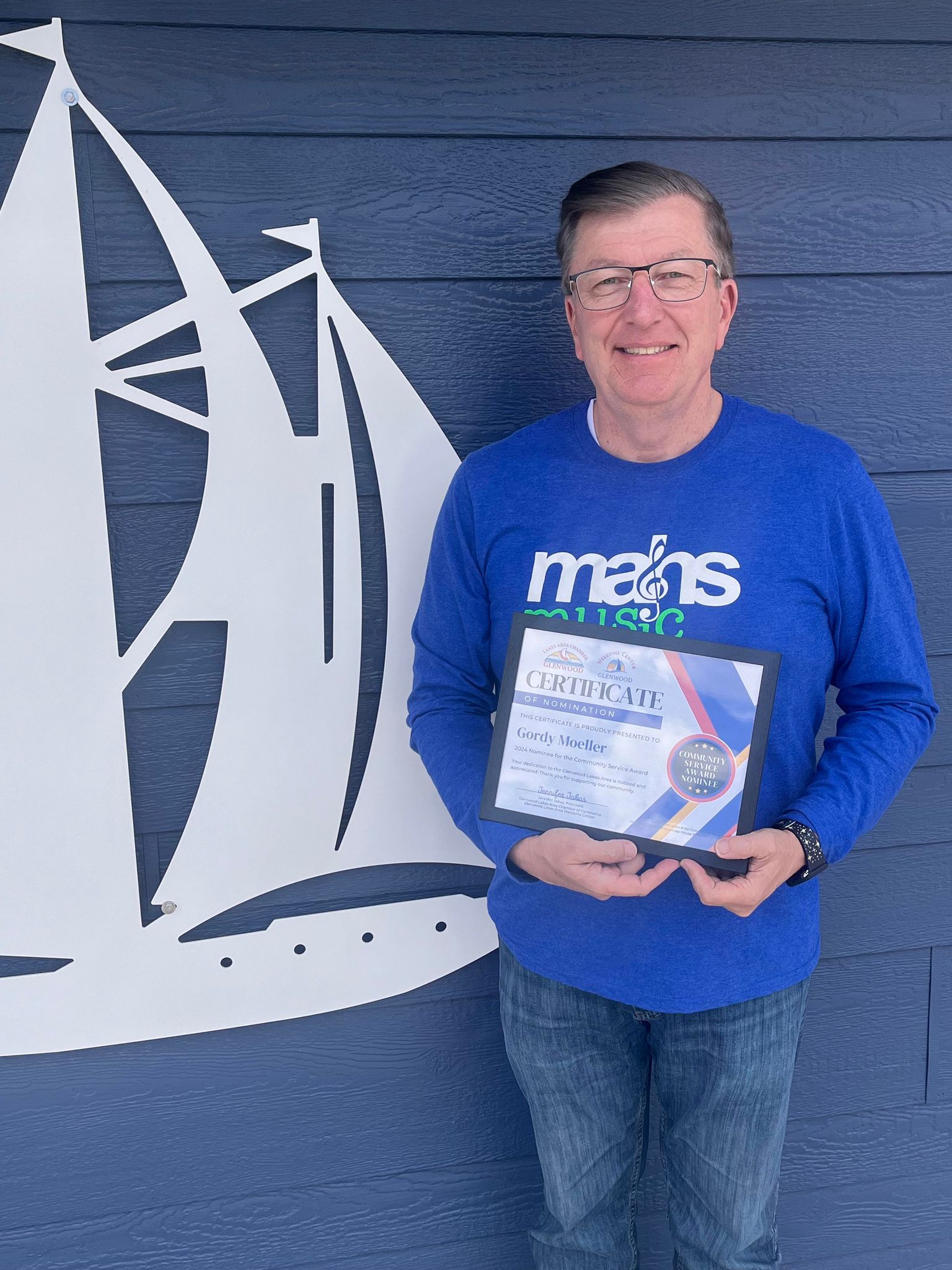 A person wearing a blue long-sleeve shirt stands against a wall with a sailboat graphic, holding a framed certificate.
