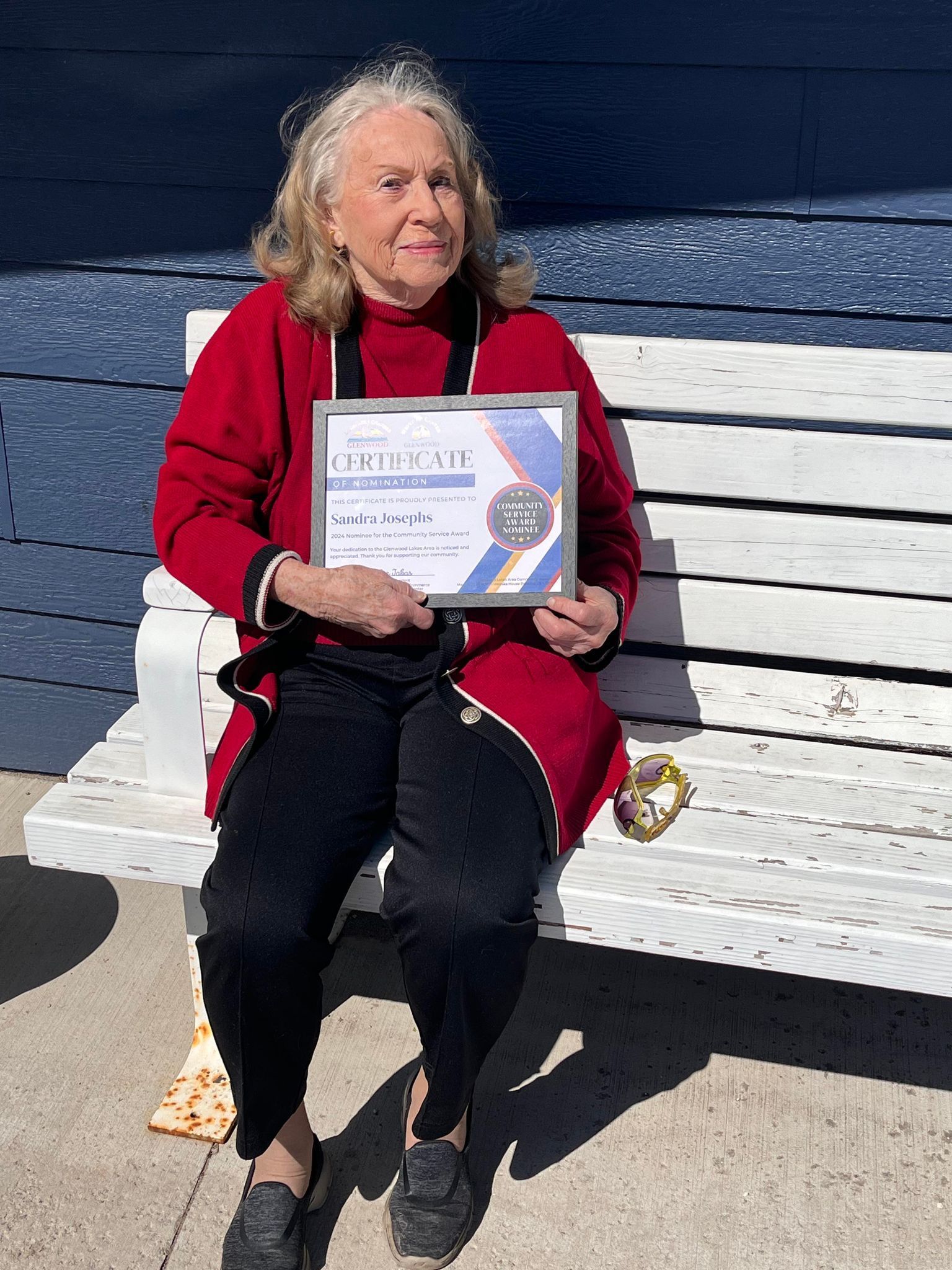 A person in a red sweater sits on a white bench outdoors, holding a framed certificate.