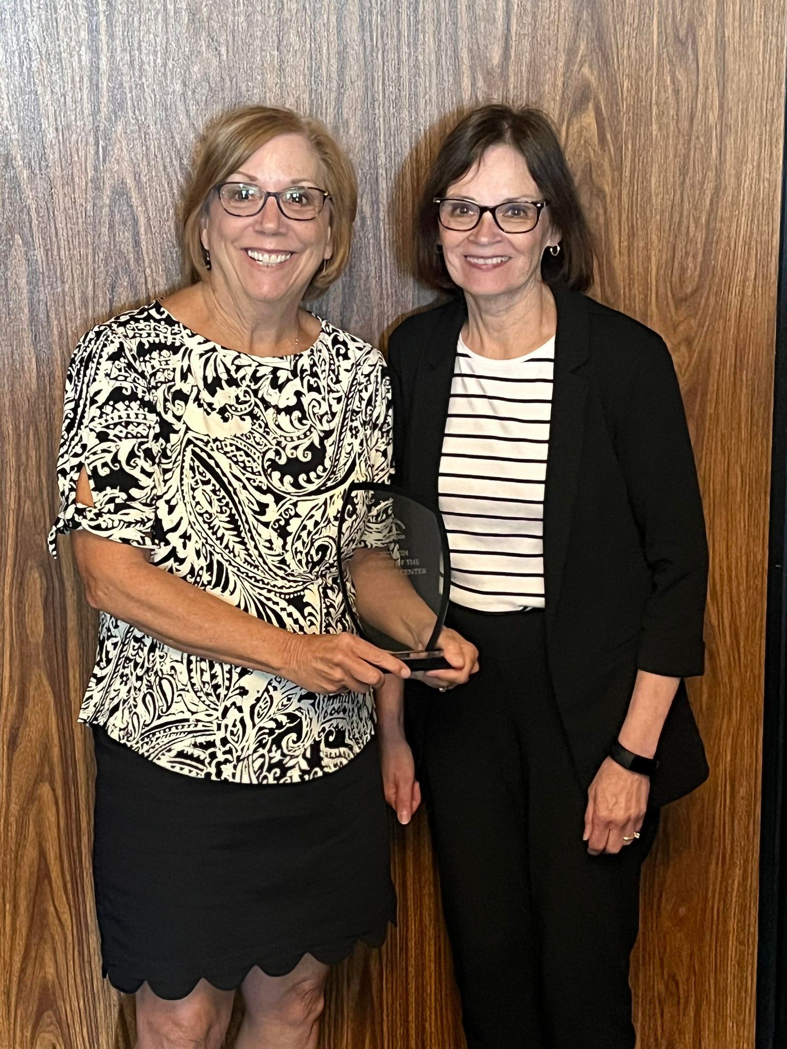 Two people stand together in front of a wood-paneled wall; one holds a glass award while the other smiles.