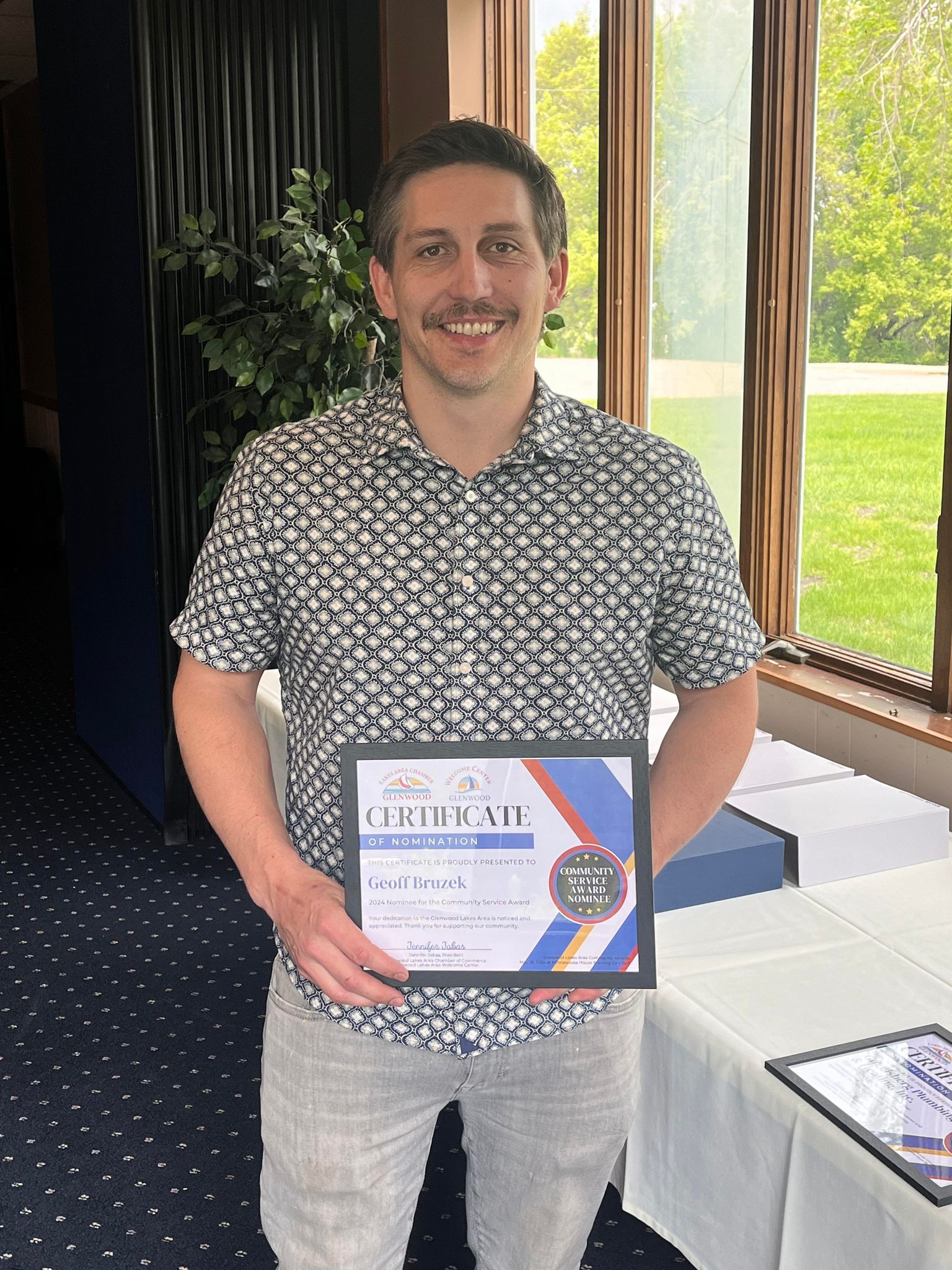 A person in a patterned shirt smiles while holding a framed certificate in a sunlit indoor setting.