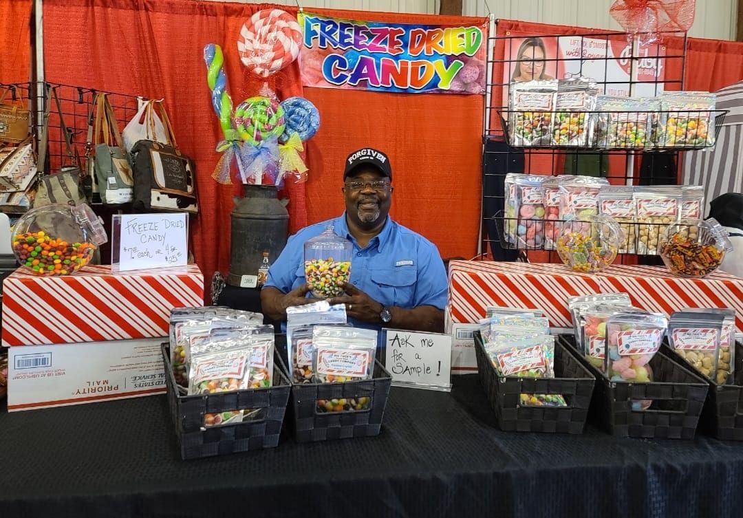 A smiling vendor in a blue shirt behind a booth selling colorful freeze-dried candy, displayed in bags and baskets.