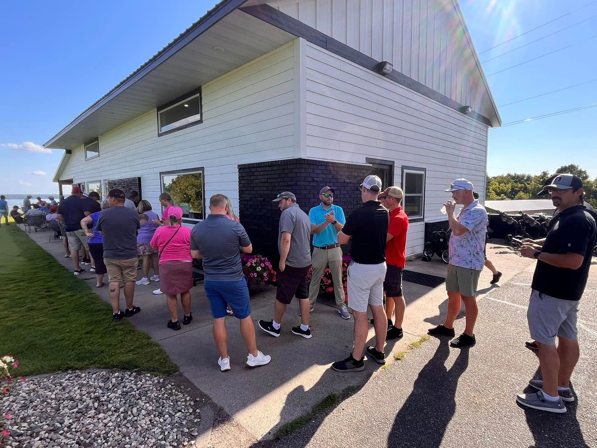 A group of people standing in a line outdoors next to a white building on a sunny day.
