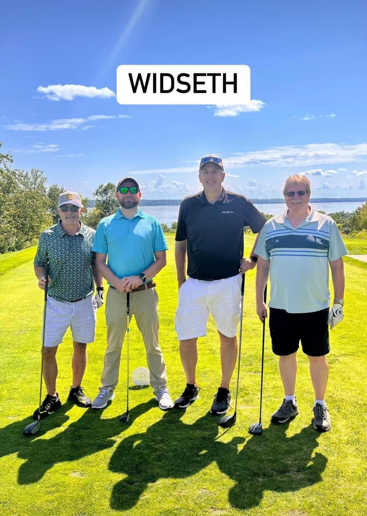 Four people pose with golf clubs on a sunny golf course with a lake in the background. A white box reads 