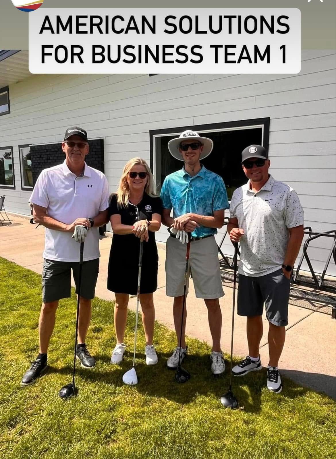 Four people stand in a line on a grassy area in front of a white building, holding golf clubs and wearing casual attire.