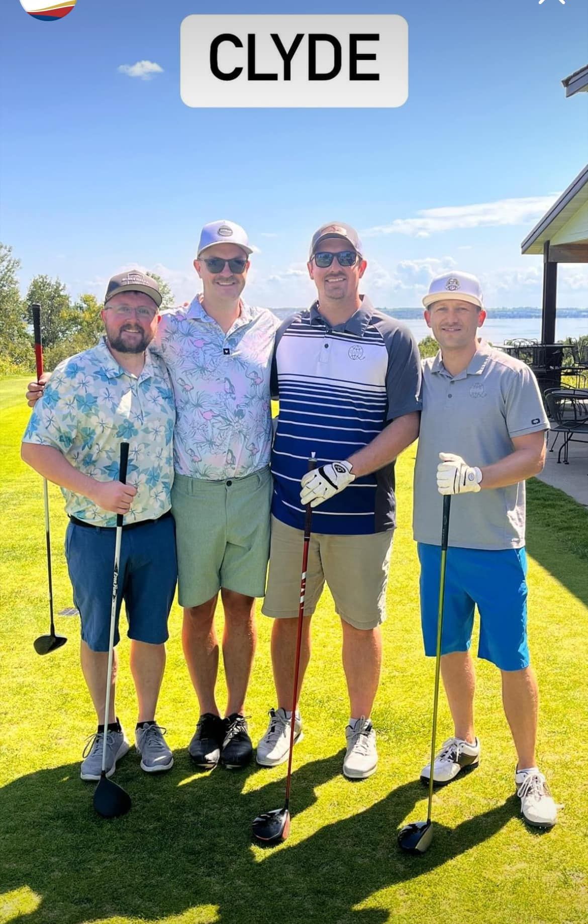 Four people in golf attire, hats, and sunglasses stand together on a grassy golf course under a blue sky.