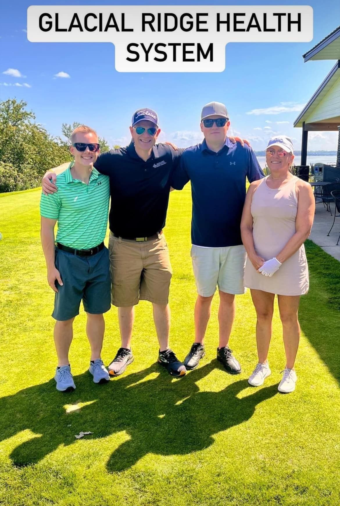 Four people stand together outdoors on a sunny, grassy lawn, smiling in golf attire under a Glacial Ridge Health System sign.