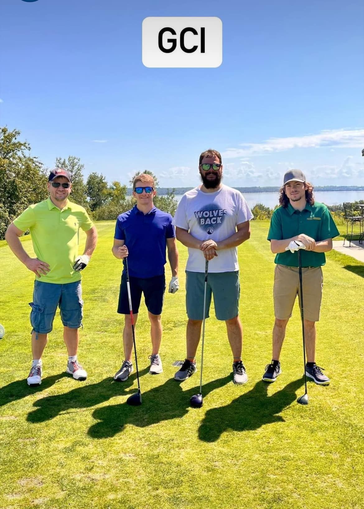 Four people standing on a sunny golf course, each holding a golf club, with a body of water in the background.