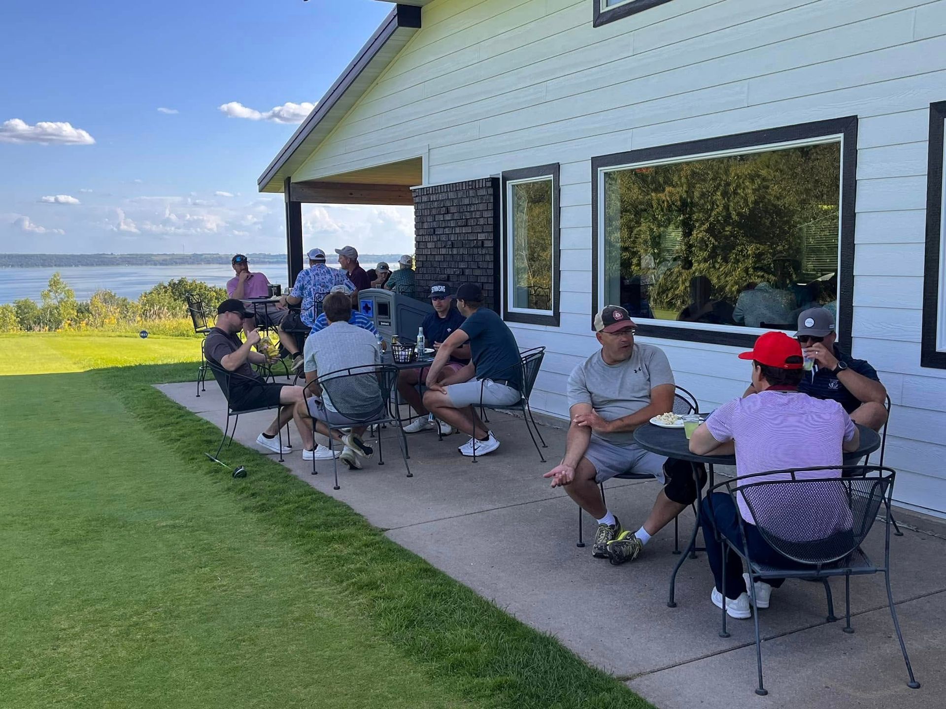 A group of people sit at patio tables outside a white building overlooking a large lake on a sunny day.