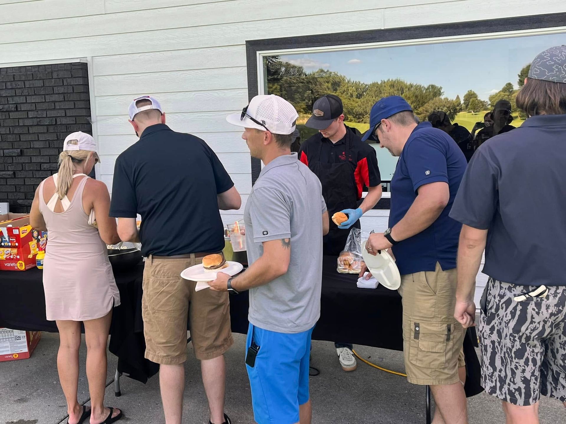 People in casual attire line up to pick up food at an outdoor buffet table in front of a white building.