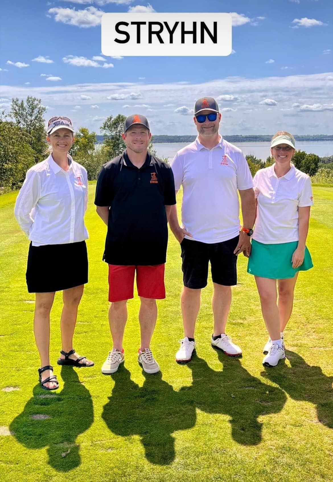 Four people stand on a grassy golf course overlooking a lake, with a 