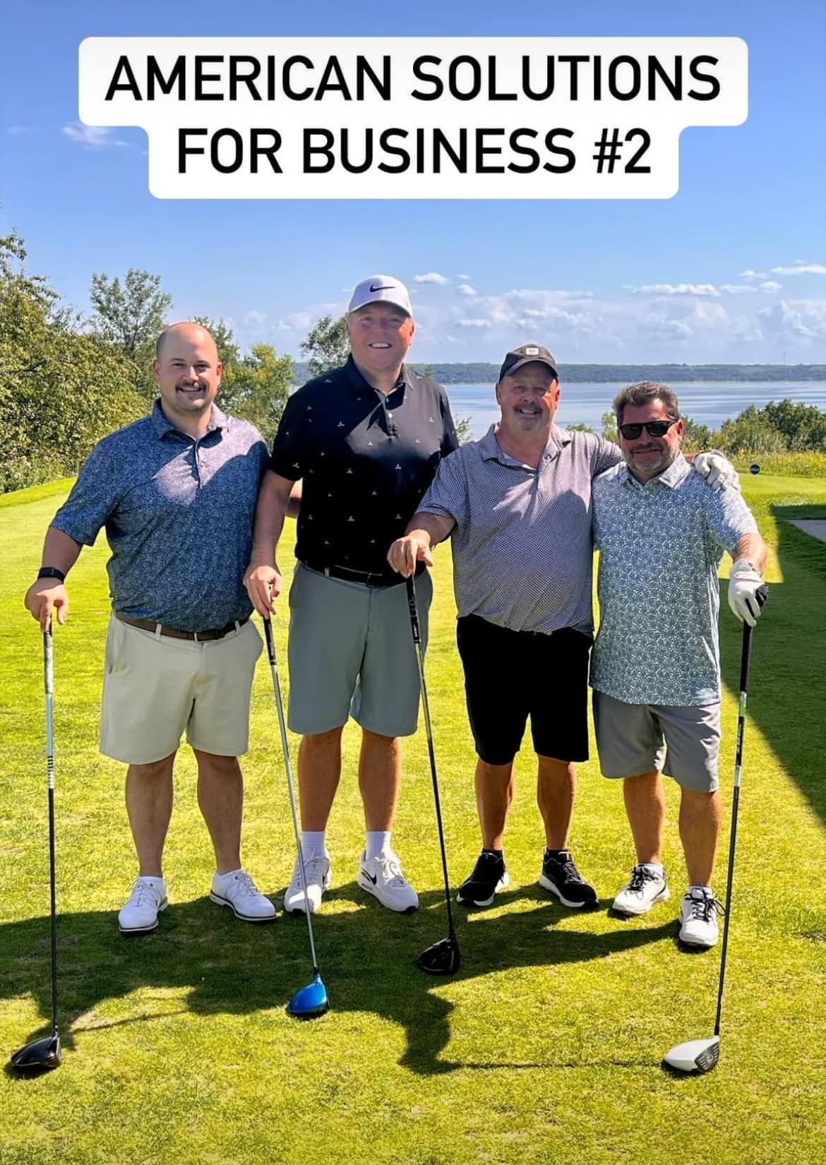 Four people in golf attire standing on a sunny golf course, holding golf clubs and posing for a group photo.