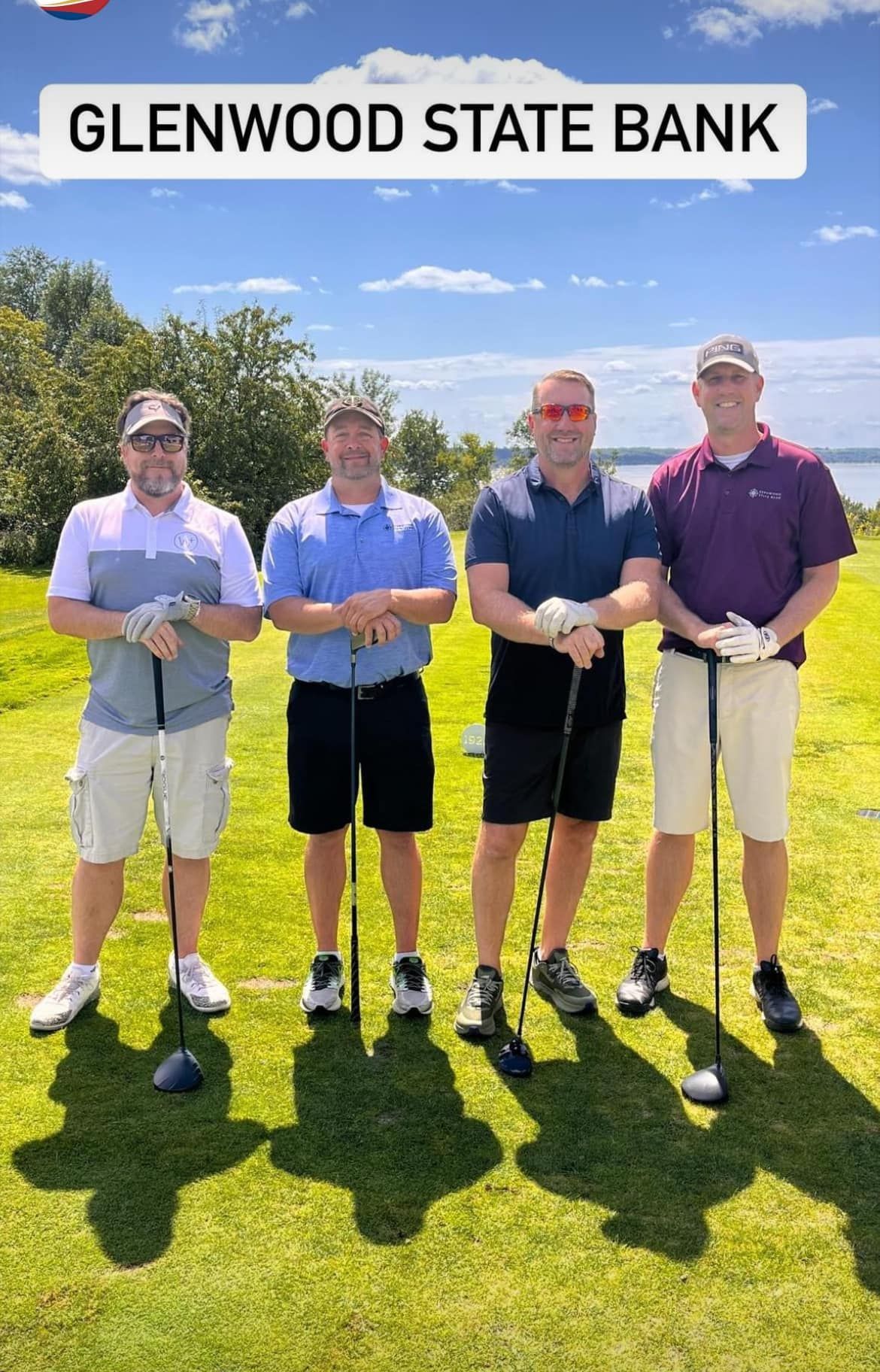 Four people in casual golf attire stand in a line on a sunny golf course, posing with golf clubs under a branded sign.