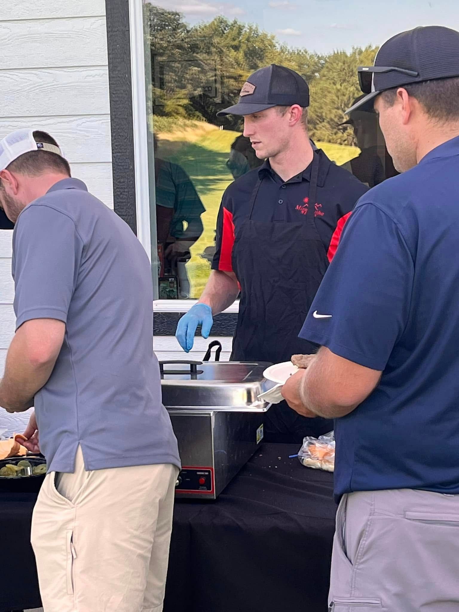 Three people stand at an outdoor table with a grill, preparing and serving food.