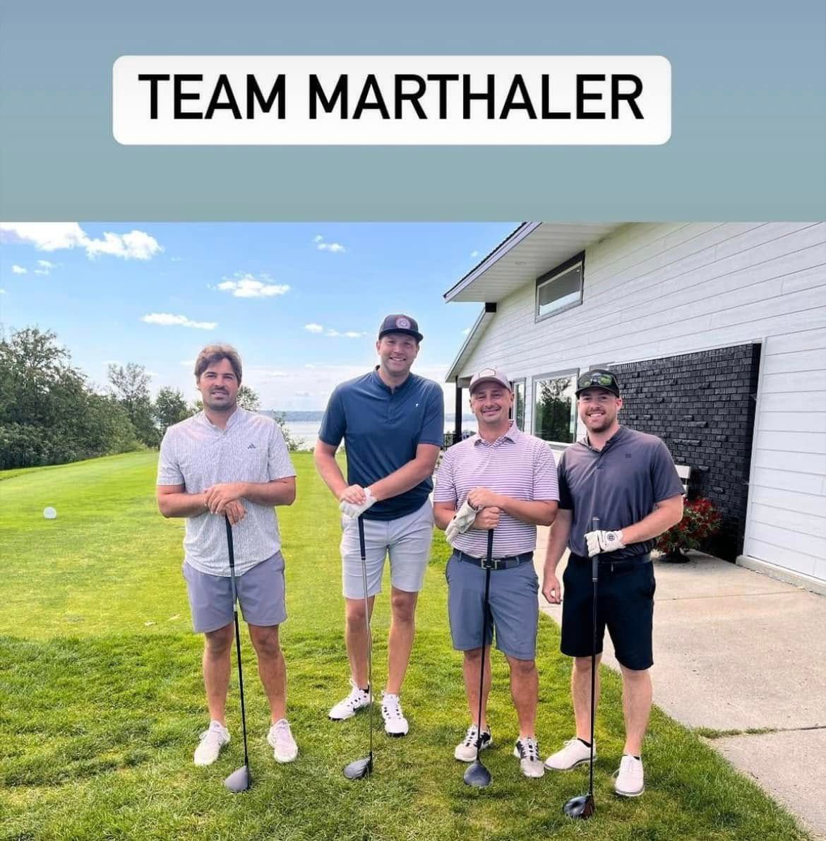 Four people in golf attire stand on a grassy lawn with golf clubs, posing in front of a house under a clear blue sky.