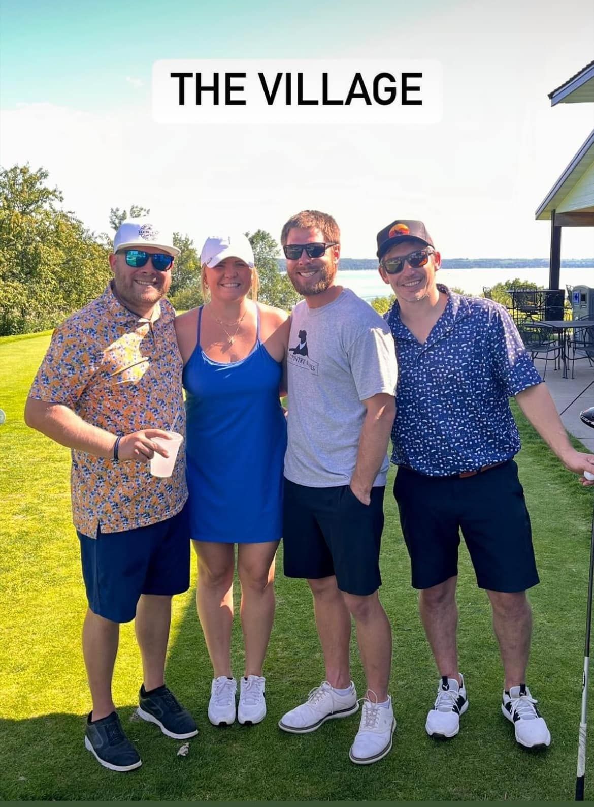 Four people stand outdoors in sunny weather, posing together on a grassy lawn with a lake in the background.