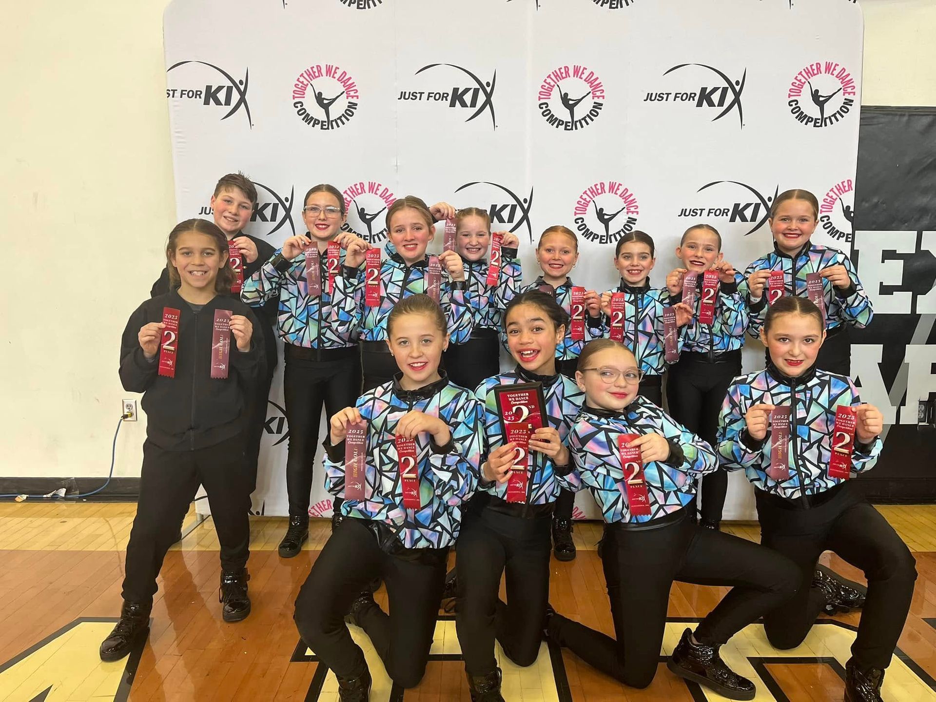 A group of twelve youth in matching patterned jackets and black pants pose with red ribbons in front of a branded banner.